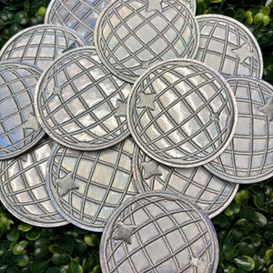 May include: A collection of silver, round patches with a globe design. Each patch features a grid pattern and a star. The patches have a holographic, iridescent finish, creating a shimmering effect. The patches are arranged on a bed of green leaves.