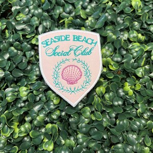May include: A pink patch with the words "SEASIDE BEACH Social Club" embroidered in teal. A teal wreath surrounds a pink seashell. The patch is on a background of green leaves.