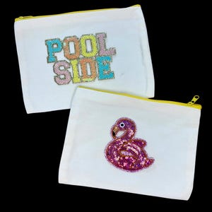 May include: Two white canvas cosmetic bags with yellow zippers. One bag has the words "POOL SIDE" in colorful sequined letters. The other bag features a pink sequined flamingo design. Both bags are on a black background.