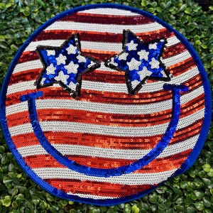 May include: Round sequined patch with a patriotic design. The patch features red and white stripes, two blue star-shaped eyes filled with white stars, and a blue smiling mouth. The border is blue.