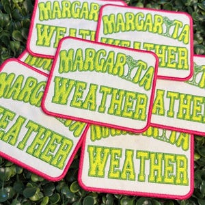 May include: A collection of embroidered patches with the words "Margarita Weather" in a lime green, bubble-style font. The patches have a white background with a hot pink border. The patches are arranged on a bed of green leaves.