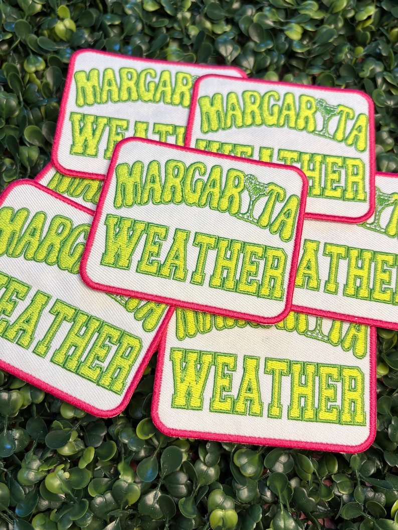 May include: A collection of embroidered patches with the words "Margarita Weather" in a lime green, bubble-style font. The patches have a white background with a hot pink border. The patches are arranged on a bed of green leaves.