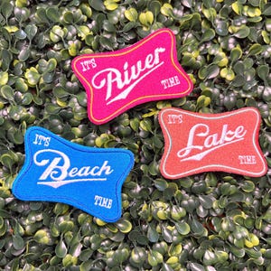 May include: Three embroidered patches with the phrases "It's River Time", "It's Beach Time", and "It's Lake Time". The patches are in pink, blue, and orange, with white lettering. The patches are on a green leafy background.