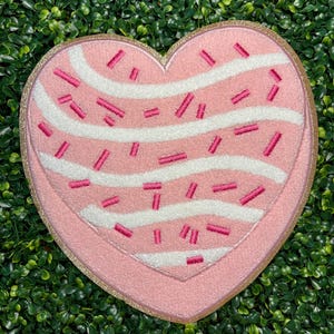 May include: A heart-shaped pink rug with white wavy stripes and pink sprinkles. The rug has a gold glitter border. The rug is on a green surface.