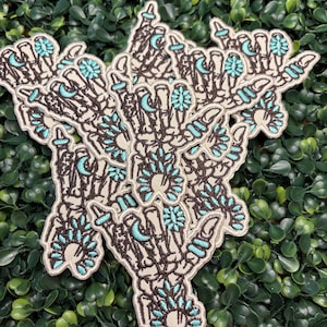 May include: Embroidered patches featuring a skeletal hand design with turquoise accents. The patches have a cream-colored background with detailed black outlines and turquoise floral and crescent moon details. The patches are suitable for jackets, bags, or other fabric items.