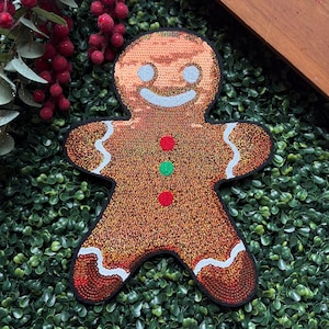 May include: A sequined gingerbread man patch with a smiling face, red, green, and red buttons, and white icing details. The patch is outlined in black and set against a green background, perfect for holiday crafts.