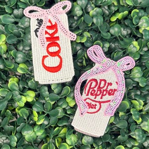 May include: Two embroidered patches shaped like soda cans, one labeled Diet Coke and the other Dr. Pepper. Each patch features a light gray background with red text and is adorned with a decorative pink sequined bow at the top of the can.