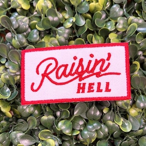 May include: A rectangular patch with a white background and a red border. The patch features the words "Raisin' HELL" in red script and block letters. The patch is set against a backdrop of green foliage.