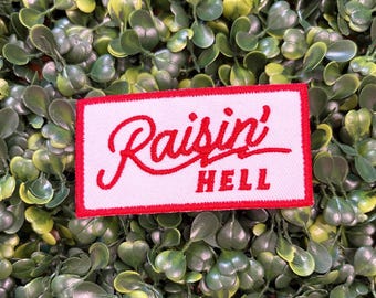 May include: A rectangular patch with a white background and a red border. The patch features the words "Raisin' HELL" in red script and block letters. The patch is set against a backdrop of green foliage.
