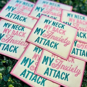 May include: A collection of square fabric patches with the embroidered text "MY NECK, MY BACK, MY Anxiety ATTACK" in pink and teal. The patches have a cream-colored background and a pink border. The patches are arranged on a bed of green leaves.