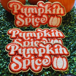 May include: Three embroidered patches featuring the text Pumpkin Spice in a bold, orange and white retro font. Each patch includes a small, stitched pumpkin icon on the right side. The patches have a gold glitter border and a textured surface.