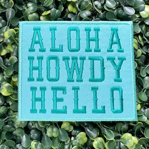 May include: A square, turquoise patch with the words "ALOHA HOWDY HELLO" embroidered in a matching teal color. The patch is surrounded by green foliage.