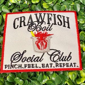 May include: Embroidered patch with a red border and the words "CRAWFISH Boil Social Club" in black script. A red crawfish is in a pot with a yellow corn cob. The text "PINCH.PEEL.EAT.REPEAT." is below.