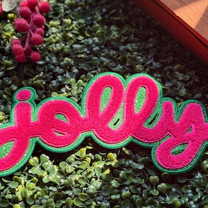 May include: A patch featuring the word jolly in bright pink cursive script with a glittery green fill, outlined by a thick green border. The lettering has a textured appearance and is designed to be applied to fabric surfaces as a decoration.
