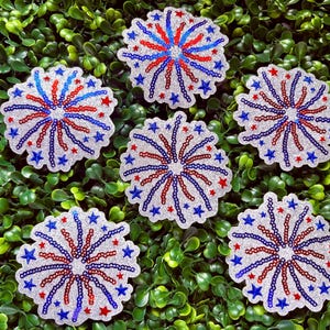 May include: Six glittery, star-shaped patches with a firework design. Each patch features red and blue lines radiating from a central point, with blue and red star accents. The patches are set against a backdrop of green foliage.