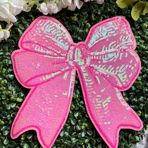 May include: A large, bright pink sequined bow with a darker pink outline. The sequins have an iridescent sheen, creating a shimmering effect. The bow is set against a backdrop of green foliage and white flowers.