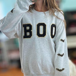 May include: A light gray crewneck sweatshirt featuring the word BOO in large black letters with gold glitter outlines on the chest. The sleeves are decorated with small black and gold glitter bat appliques arranged in a vertical pattern.