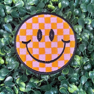May include: A round patch featuring a smiley face design. The face has a checkerboard pattern in orange and pink, with black eyes and a curved smile. The patch is outlined in black and set against a background of green leaves.
