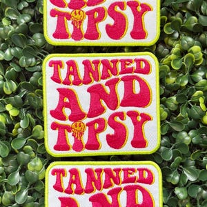 May include: Three embroidered patches with the words "TANNED AND TIPSY" in pink, outlined in yellow, on a white background. The patches have a yellow border and a smiling face graphic. The patches are displayed on a green leafy background.