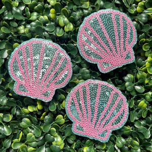 May include: Three seashell-shaped patches with pink sequin details and teal sequin filling. The patches are arranged on a bed of green foliage, creating a vibrant and eye-catching display.