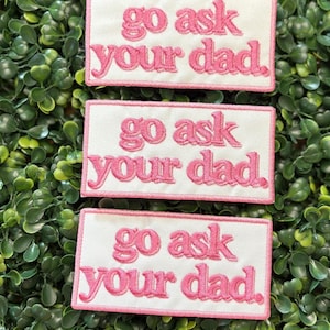 May include: Three rectangular patches with the phrase "go ask your dad" embroidered in pink on a white background. The patches are arranged on a bed of green foliage.