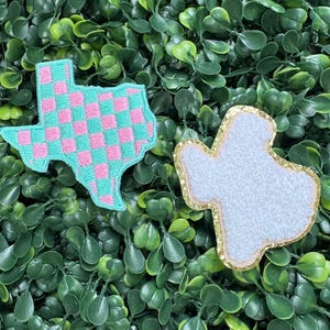 May include: Two Texas-shaped patches. One is teal with a pink checkered pattern. The other is white with a gold glitter border. Both are set against a background of green foliage.