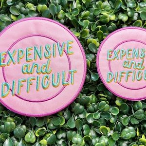 May include: Two pink embroidered patches with the text "EXPENSIVE and DIFFICULT" in teal and yellow. The patches are circular with a pink border and are set against a backdrop of green foliage.