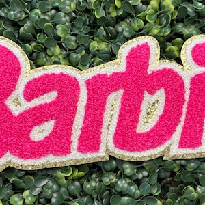 May include: A pink and white embroidered patch with the name "Barbie" in a cursive font. The patch has a gold glitter border and is set against a green leafy background. The patch is a decorative item.
