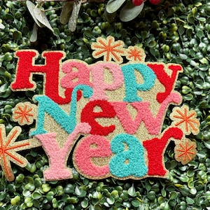 May include: A colorful Happy New Year sign with red, pink, and teal letters on a gold glitter background. The sign is decorated with gold glitter starbursts and is set against a backdrop of green foliage.