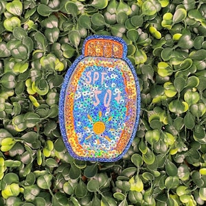 May include: A blue, orange, and gold sequined patch in the shape of a sunscreen bottle. The patch has the text "SPF 50" and a sun graphic. The patch is set against a background of green leaves.