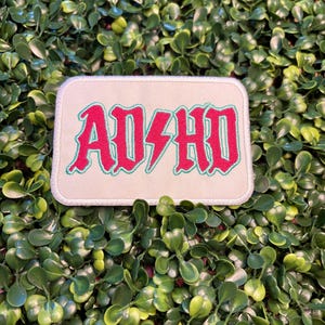 May include: A rectangular patch with the text "AD/HD" in red, outlined in green, on a cream background. The patch has a white border and is set against a backdrop of green foliage.
