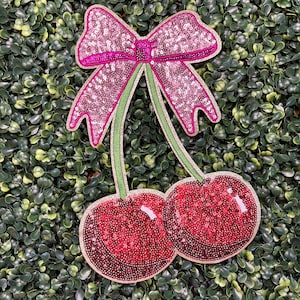 May include: A decorative applique featuring a pink sequined bow and two red sequined cherries with green stems. The applique is set against a backdrop of green foliage.