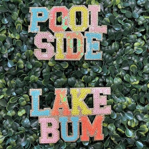 May include: Two sequined patches with the phrases "POOL SIDE" and "LAKE BUM" in colorful letters. The letters are outlined in gold and filled with sequins in shades of pink, yellow, orange, and blue. The background is a dense arrangement of green leaves.