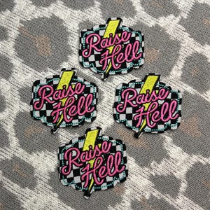 May include: Four embroidered patches with the phrase "Raise Hell" in pink script, outlined in black, with a yellow lightning bolt. The patches have a black and white checkered background and a teal border.