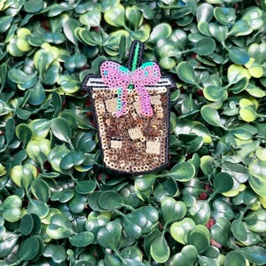 May include: A sequined patch in the shape of an iced coffee drink. The patch features brown sequins for the drink, a pink and purple bow, and a green straw. The patch is outlined in black and sits on a bed of green leaves.