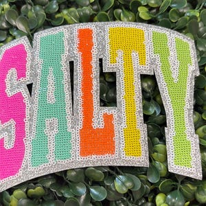 May include: A colorful patch with the word "SALTY" in a block letter design. The letters are filled with pink, teal, orange, yellow, and green sequins, outlined with silver glitter. The patch is set against a backdrop of green foliage.