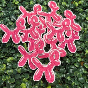 May include: A collection of pink balloon dog patches with white outlines, arranged on a bed of green foliage. The patches are embroidered with a playful design, perfect for adding a whimsical touch to clothing or accessories.