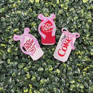 May include: Three embroidered patches featuring soda cans. One is red with "Dr Pepper" text, another is silver with "Dr Pepper" text, and the third is white with "Diet Coke" text. Each patch has a pink sequined bow.