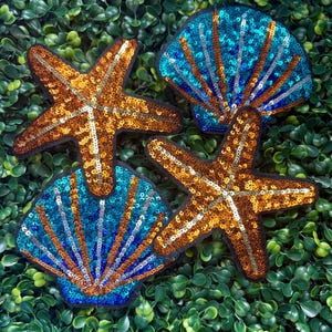 May include: Four sequined patches in the shape of seashells and starfish. Two starfish are gold and brown, and two seashells are blue and brown. The patches are on a green leafy background.