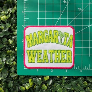May include: A rectangular patch with the words "Margarita Weather" in lime green, outlined in white, and a pink border. A lime green margarita glass graphic is above the word "Weather". The patch is on a green cutting mat.