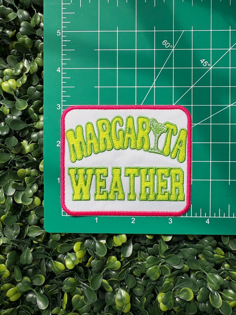May include: A rectangular patch with the words "Margarita Weather" in lime green, outlined in white, and a pink border. A lime green margarita glass graphic is above the word "Weather". The patch is on a green cutting mat.
