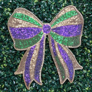 May include: A large, decorative bow with gold, green, and purple sequins. The bow is set against a backdrop of green foliage. The bow's design features alternating stripes of gold, green, and purple, creating a festive and eye-catching appearance.