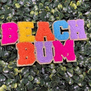 May include: Colorful, textured letters spell out "BEACH BUM" with gold glitter trim. The letters are in shades of pink, yellow, orange, blue, and purple, set against a backdrop of green foliage.