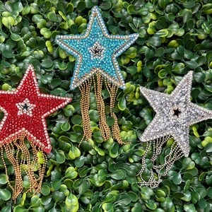 May include: Three star-shaped brooches with rhinestone embellishments and dangling chain fringe. One is red, one is blue, and one is silver. Each has a smaller star in the center. The brooches are set against a green leafy background.