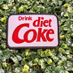 May include: A rectangular patch with the words "Drink diet Coke" in red lettering on a white background. The patch has a black border and is set against a backdrop of green foliage.