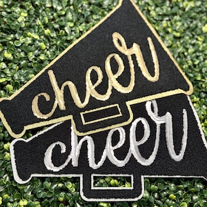 May include: Two black megaphone-shaped patches with the word "cheer" embroidered in gold and silver thread. The patches have a gold and silver border, respectively. The background is a bed of green foliage.