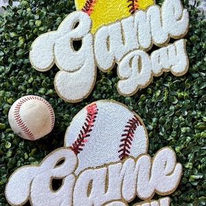 May include: A decorative arrangement featuring a baseball and a softball, each with a sequined design. The words "Game Day" are displayed in a white, textured font with gold trim. The background is a bed of green foliage.
