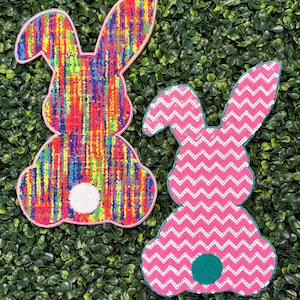 May include: Two Easter bunny-shaped decorations. One is a vibrant rainbow pattern with a white fluffy tail and pink trim. The other is pink with a white zigzag pattern, a teal tail, and teal trim. Both are set against a green leafy background.