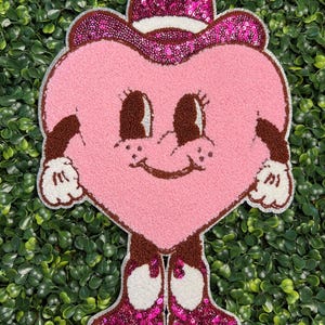 May include: A pink heart-shaped character with a cowboy hat and boots. The hat and boots are embellished with pink sequins. The character has a smiling face, brown arms, and white gloves. The background is a green hedge.
