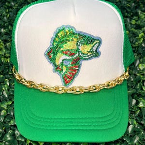 May include: A green and white trucker hat with a gold chain and a fish design. The hat features a green brim and mesh back, with a white front panel. The fish is embroidered with green, yellow, red, and blue sequins.